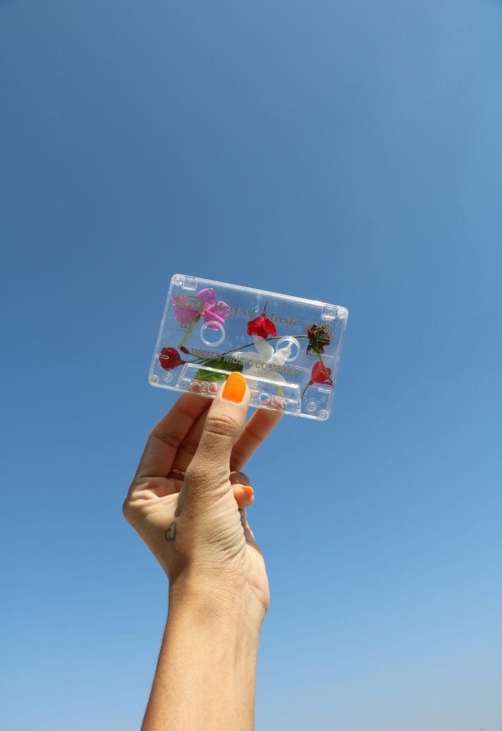 A hand with painted nails holds a flower-decorated cassette against a bright clear blue sky, symbolizing nostalgia and creativity.